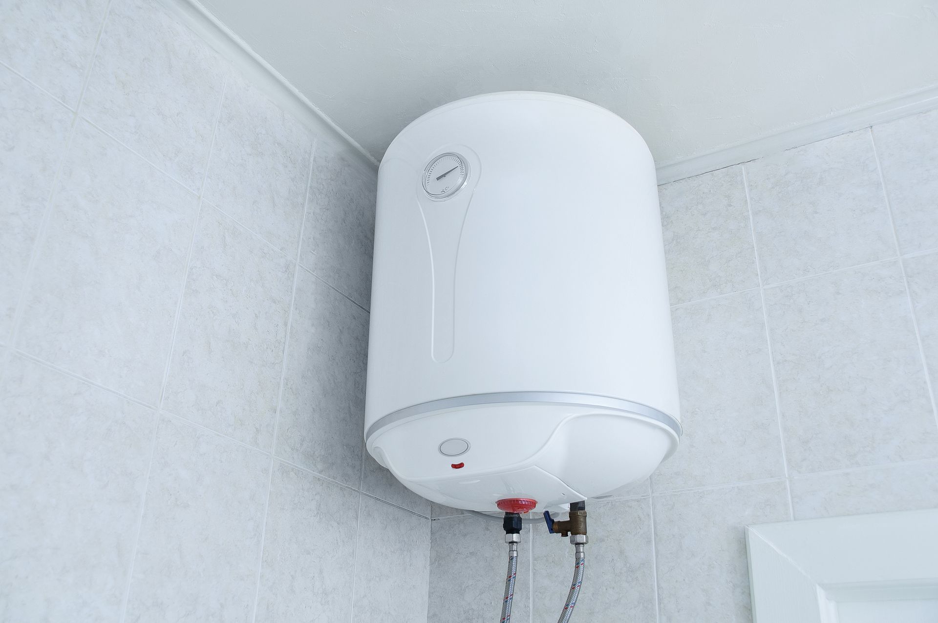 Chauffe-eau mural blanc installé dans un coin carrelé d'une salle de bain, avec les tuyaux en dessous.
