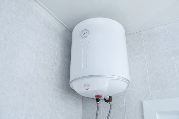 Chauffe-eau mural blanc installé dans un coin carrelé, avec tuyaux et vannes en dessous.