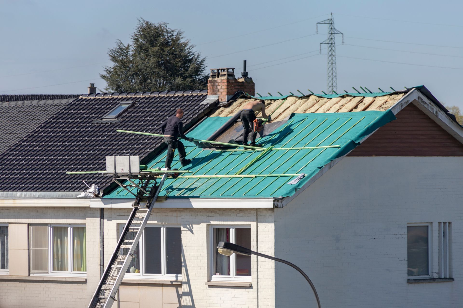 Des ouvriers refont la toiture d'une maison avec des panneaux métalliques verts à côté d'un toit sombre existant.