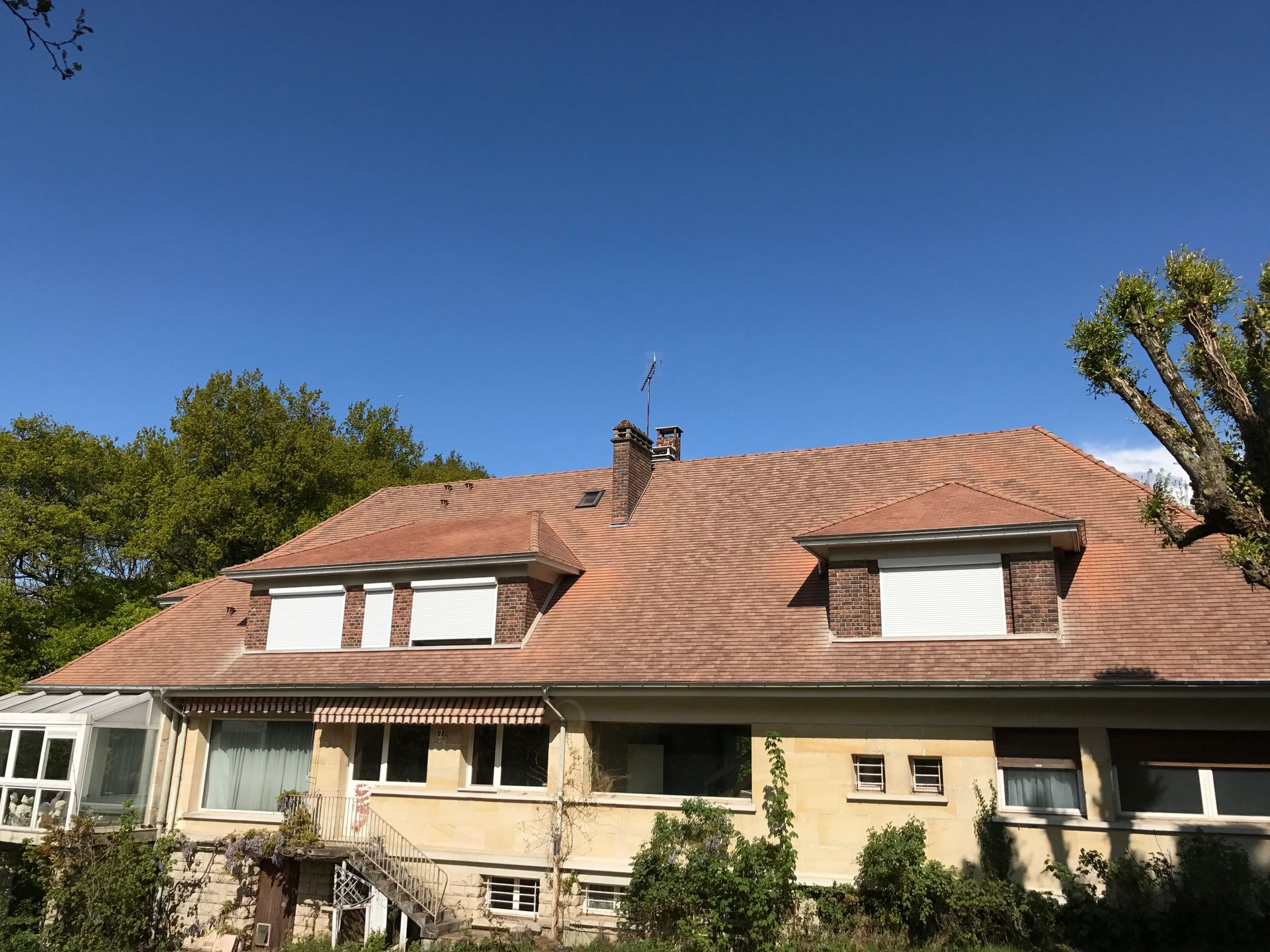 Maison beige au toit de tuiles et lucarnes sous un ciel bleu clair.