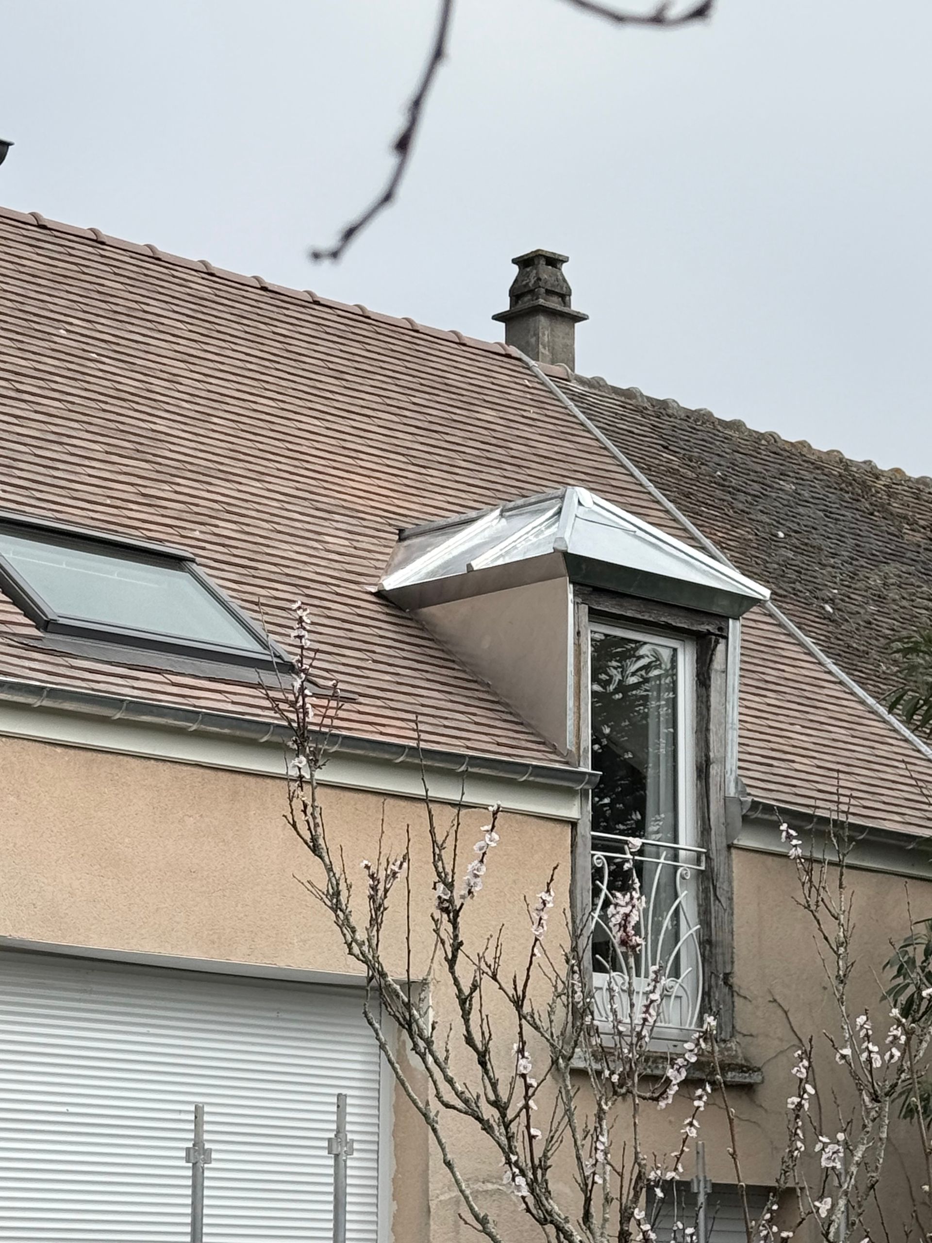 Maison avec toit de tuiles et petite lucarne, partiellement masquée par des branches d'arbres.