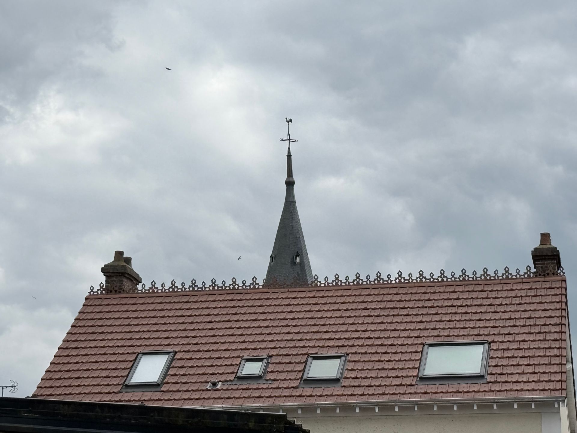 Toiture en tuiles avec trois puits de lumière et clocher d'église se détachant sur un ciel nuageux.