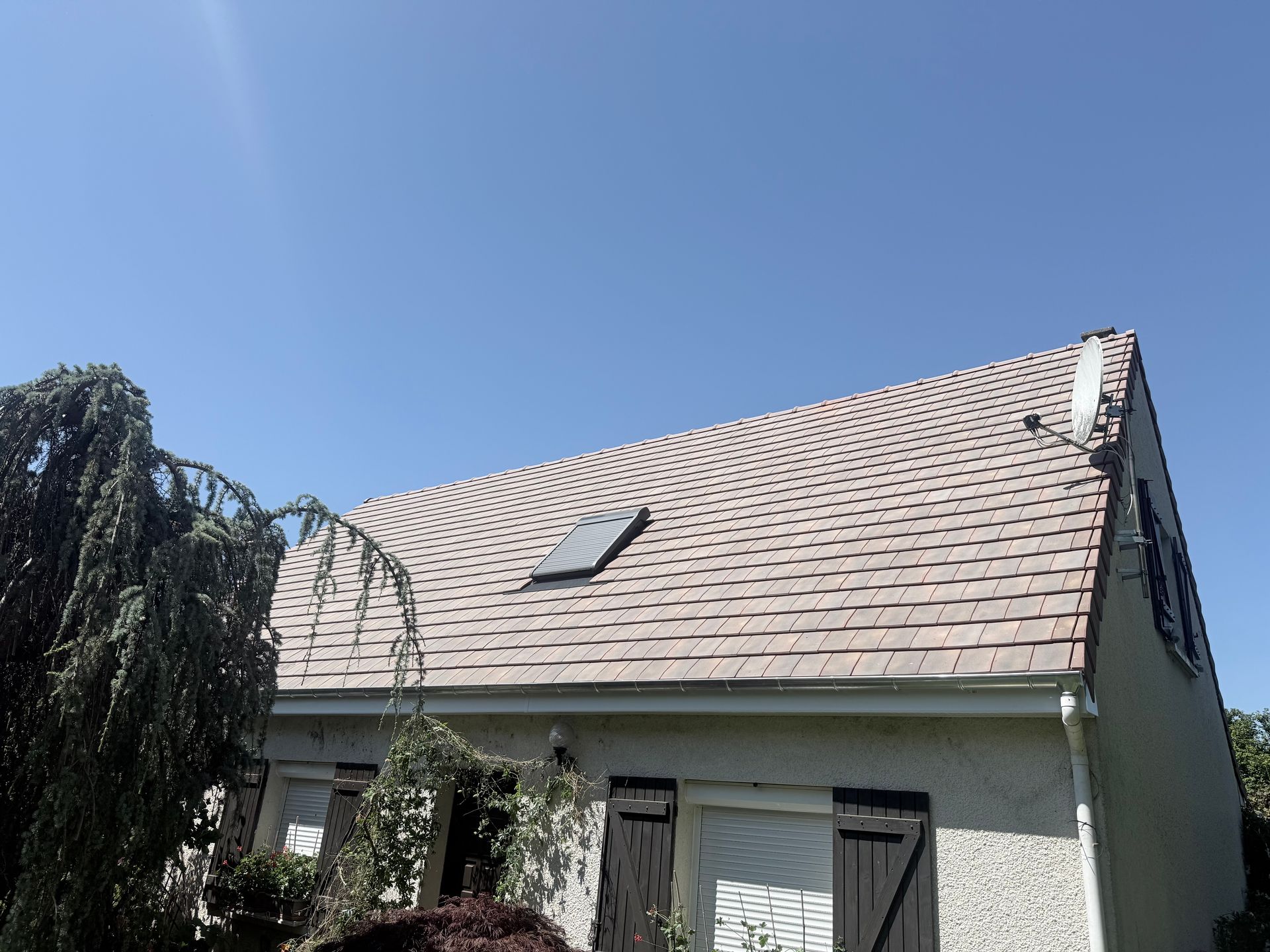 Maison avec toit pentu en bardeaux et puits de lumière sous un ciel bleu clair.