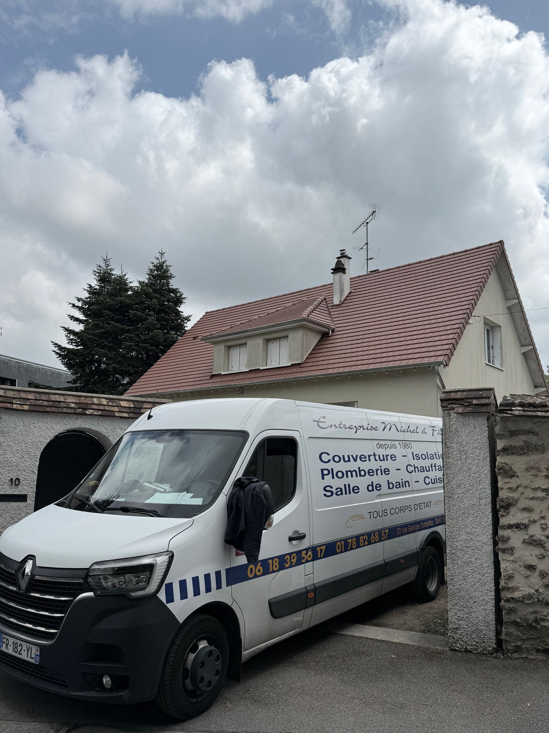 Camionnette de livraison blanche garée contre un muret en pierre devant une maison, sous un ciel nuageux.