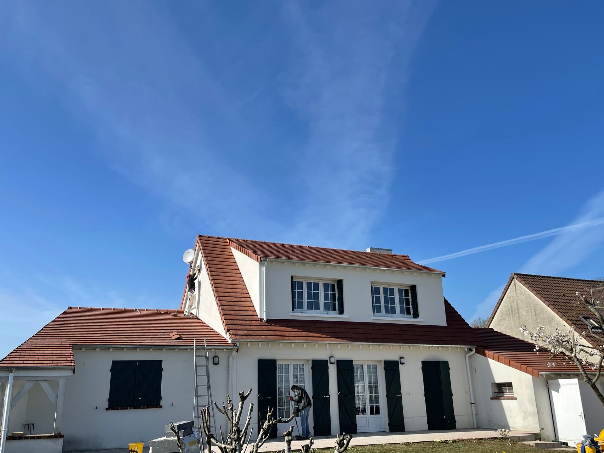 Maison blanche au toit de tuiles rouges, aux volets bleus et à la terrasse avant sous un ciel d'un bleu limpide.