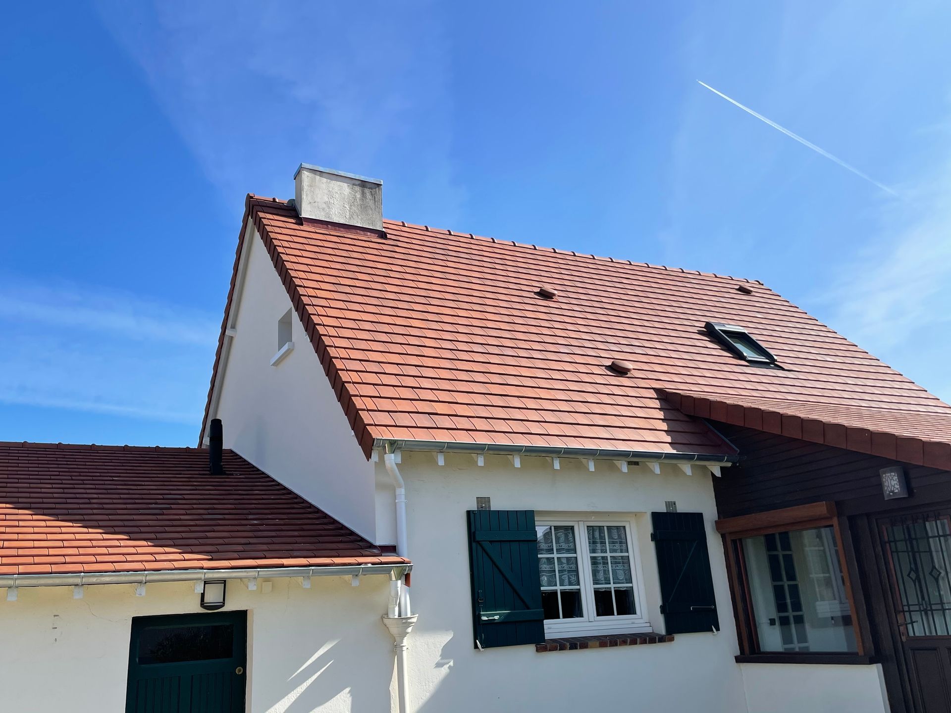 Maison blanche au toit de tuiles rouges, aux volets verts et aux puits de lumière, sous un ciel d'un bleu éclatant.