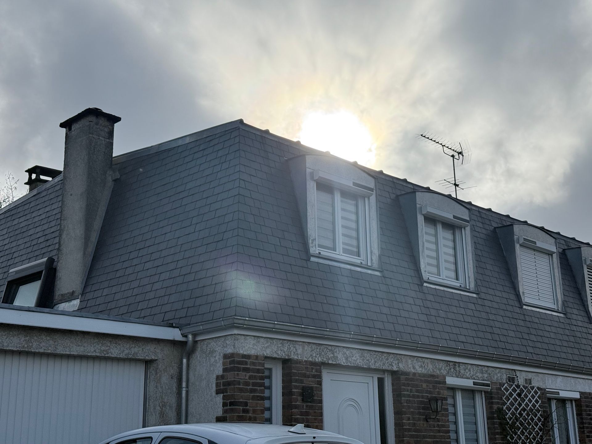 Maison au toit de bardeaux gris et à la façade en briques, le soleil perçant un ciel nuageux derrière elle.