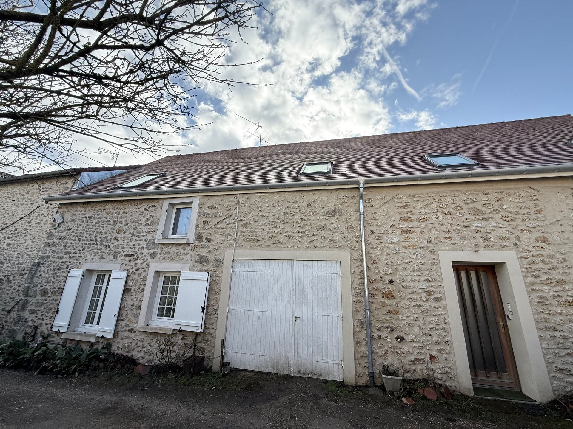 Maison en pierre avec volets blancs et porte de garage sous un toit de tuiles, vue de l'extérieur.