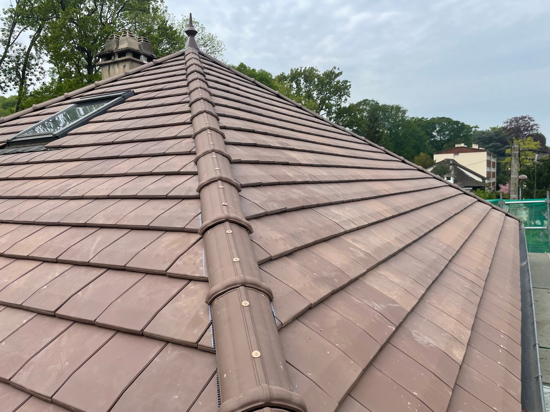 Toit en tuiles brunes avec une ventilation centrale au faîte, vu du ciel et des arbres.