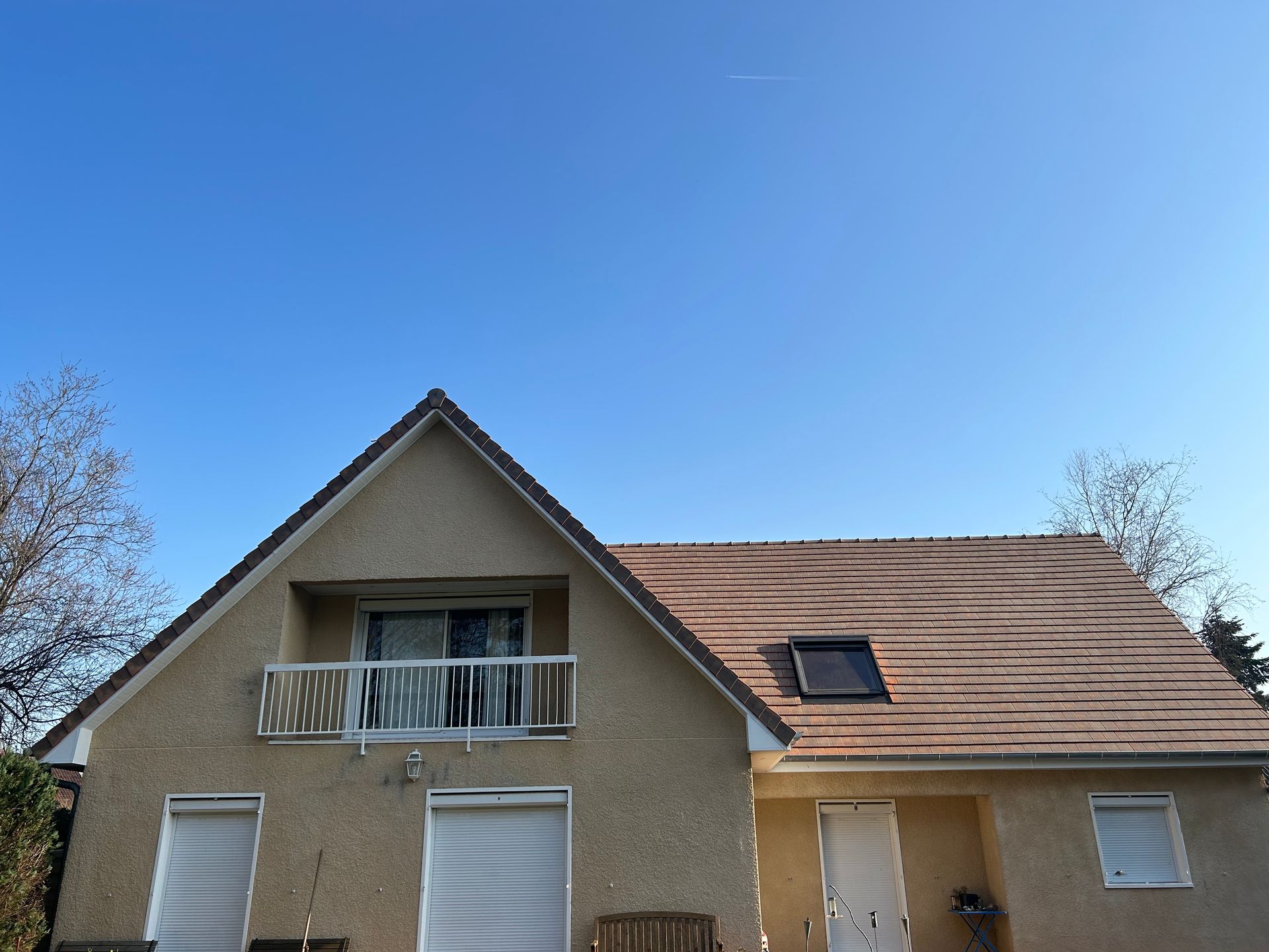 Maison à façade beige, toit de tuiles brunes, balcon et volets blancs sous un ciel bleu clair.