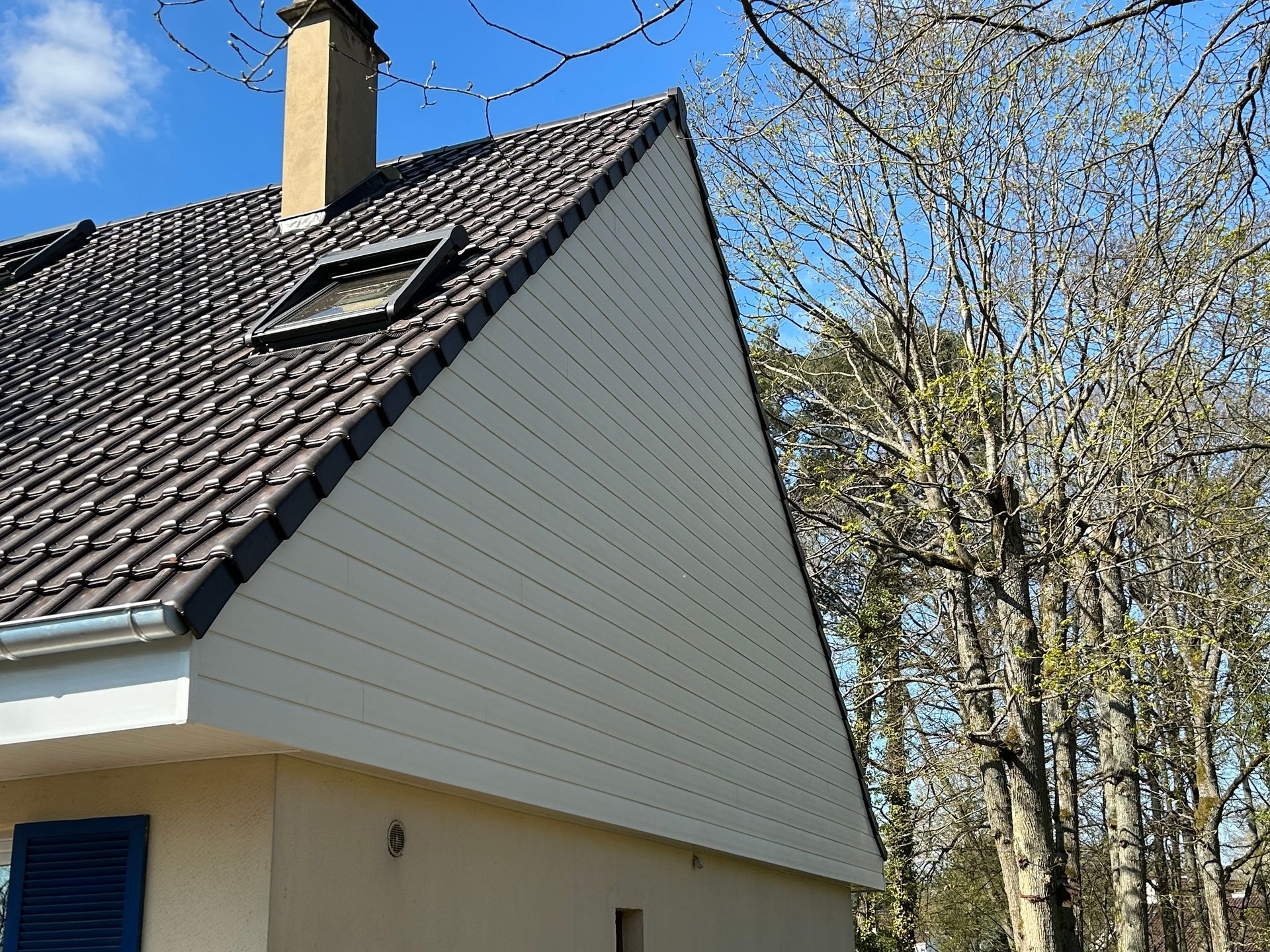 Toit de maison en bardeaux foncés, puits de lumière et cheminée se détachant sur un ciel bleu et des arbres dénudés.