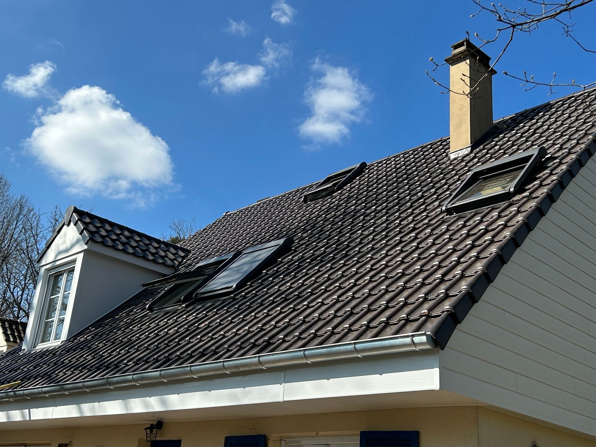 Toit de maison en tuiles sombres, fenêtres de toit, cheminée et ciel bleu nuageux.