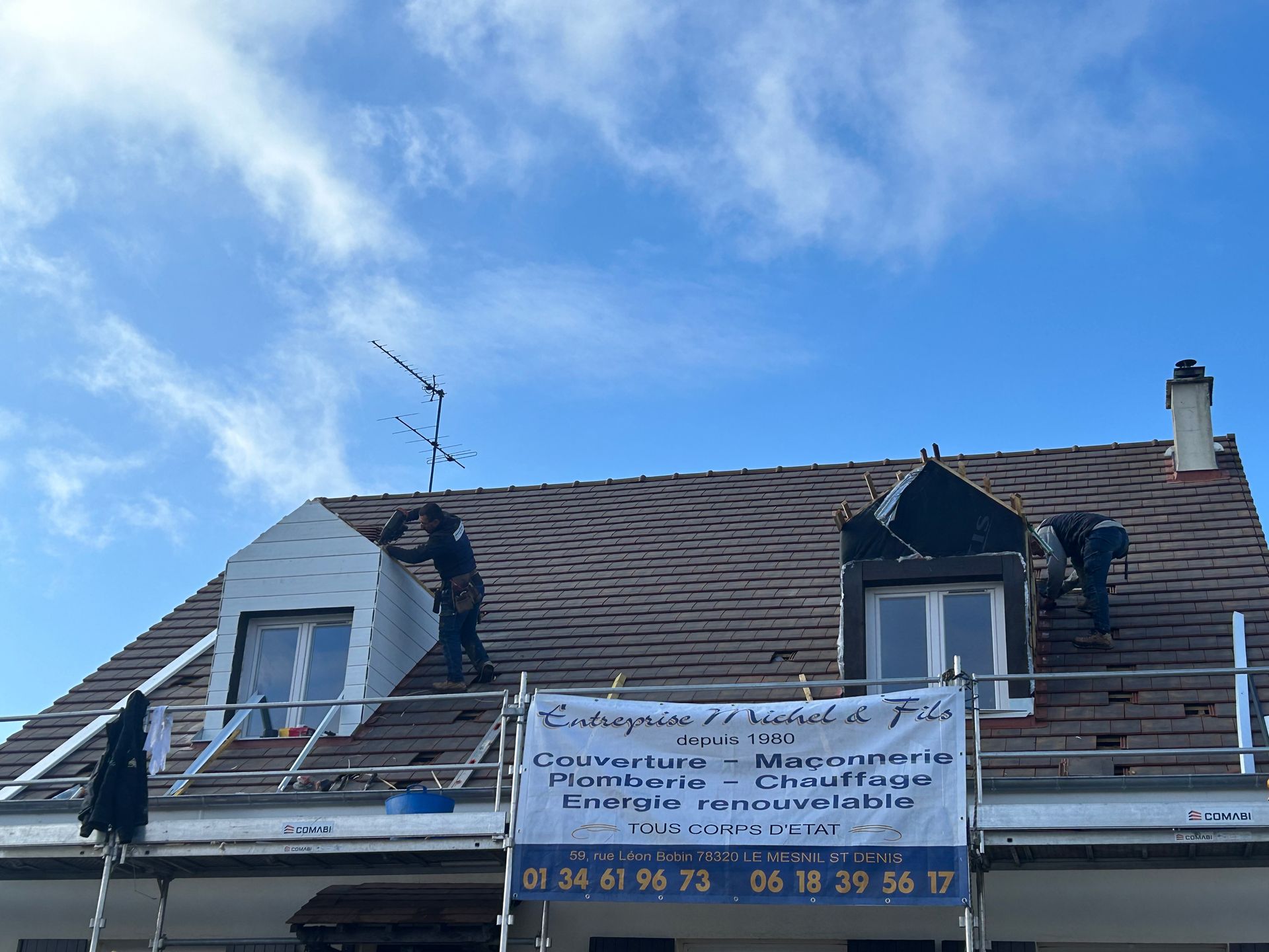 Toit de maison orné d'une grande banderole et d'un ciel bleu, montrant des dégâts visibles sur la toiture et des travaux de réparation.