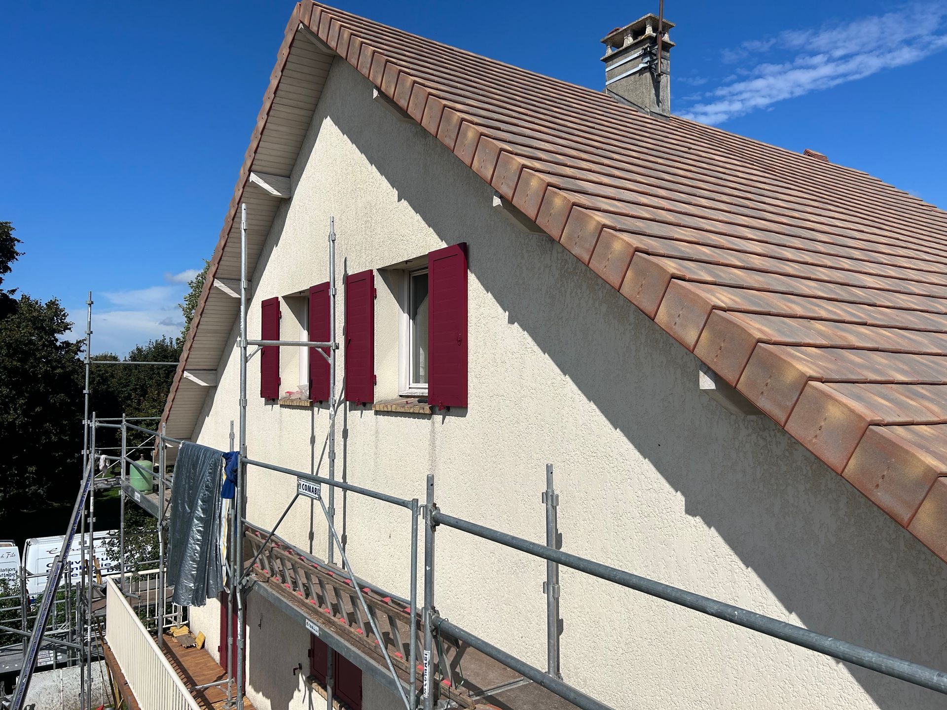 Toiture d'une maison en construction, avec des échafaudages et des fenêtres aux encadrements rouges se détachant sur un ciel bleu.