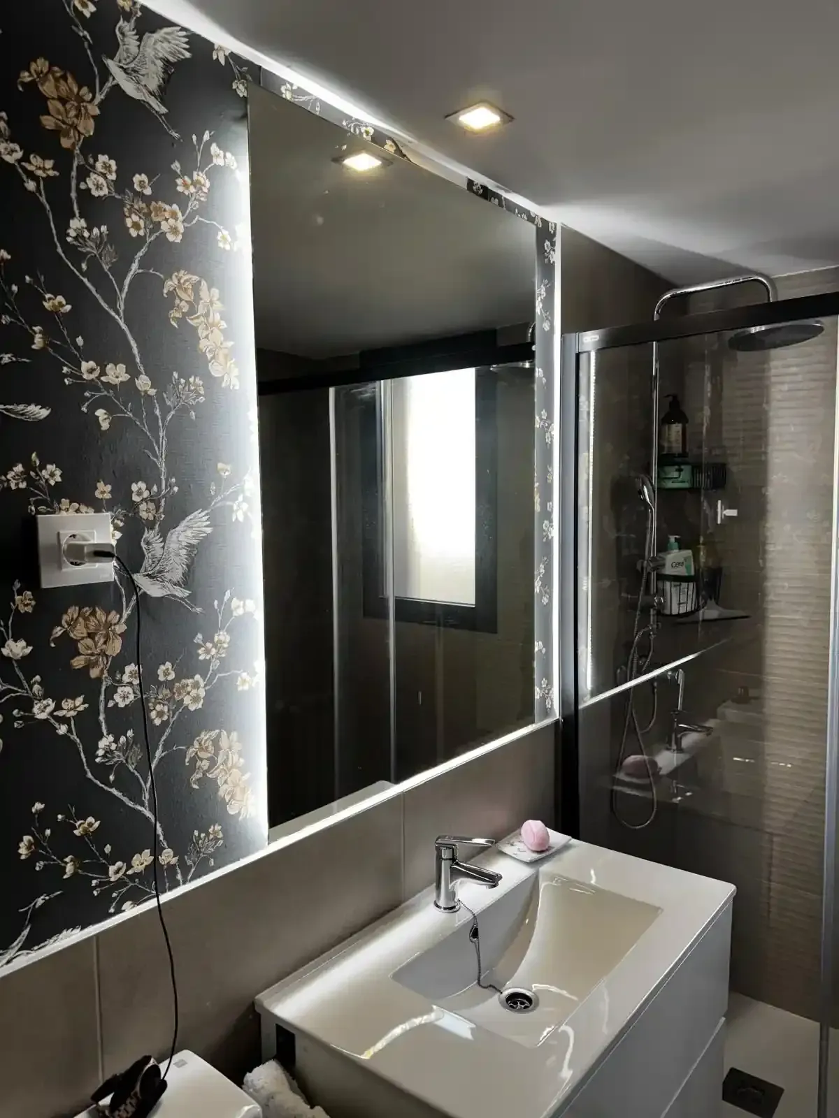 Baño con espejo iluminado, lavabo, ducha y papel pintado floral. Colores neutros con diseño moderno.