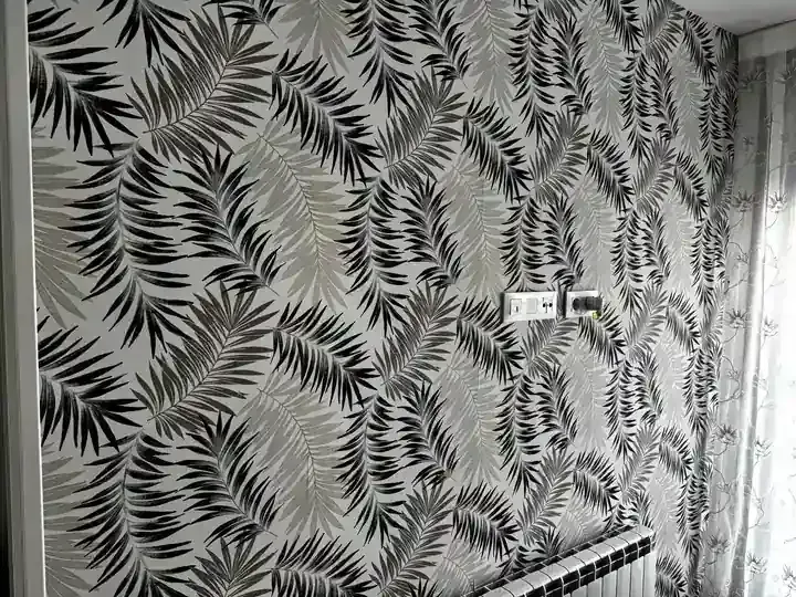 Pared con papel pintado con estampado de hojas de palmera en negro, gris y blanco. Se ven el marco de la puerta, la cortina y el radiador blancos.