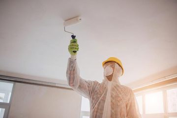 Una persona con equipo de protección pinta un techo con un rodillo en una habitación luminosa.