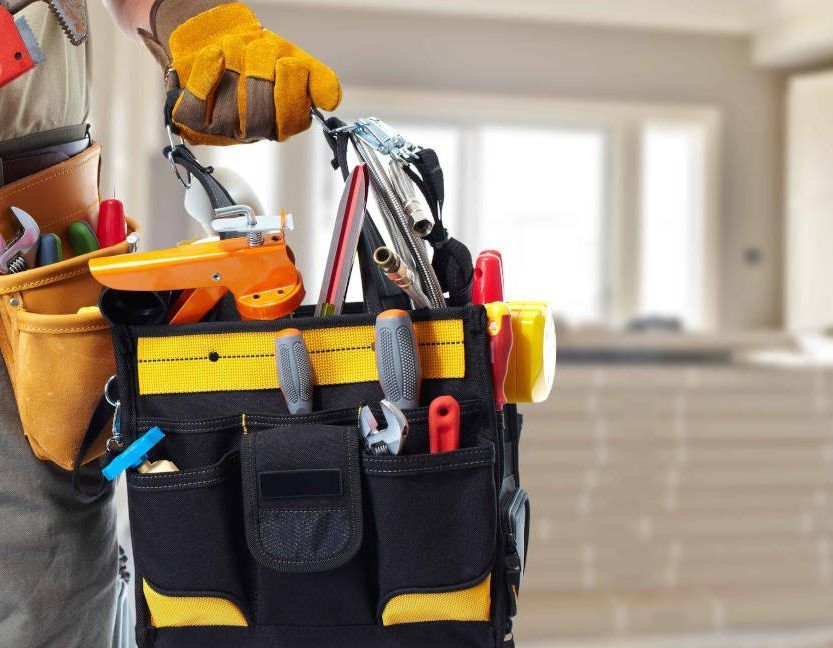 Manitas con guantes amarillos y cinturón de herramientas, sostiene herramientas con un fondo de cocina borroso.