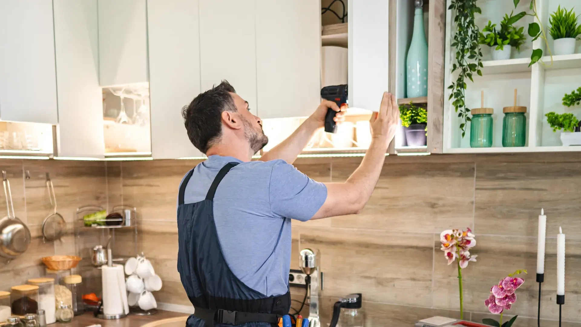 Hombre arreglando un armario en la cocina con un taladro. La cocina tiene plantas, armarios blancos y accesorios.