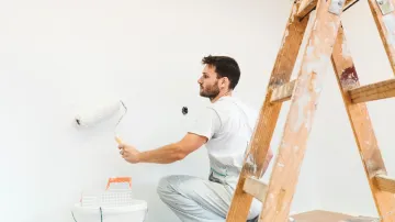 Hombre pintando una pared blanca con un rodillo de pintura, arrodillado cerca de una escalera de madera.