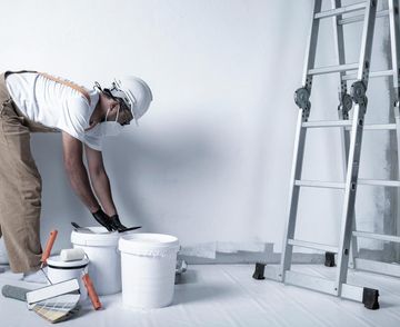 Un trabajador pinta una pared blanca junto a cubos y una escalera en una habitación.