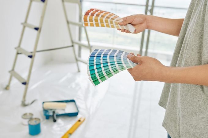 Manos sosteniendo muestras de color de pintura en una habitación luminosa con una escalera y materiales de pintura al fondo.