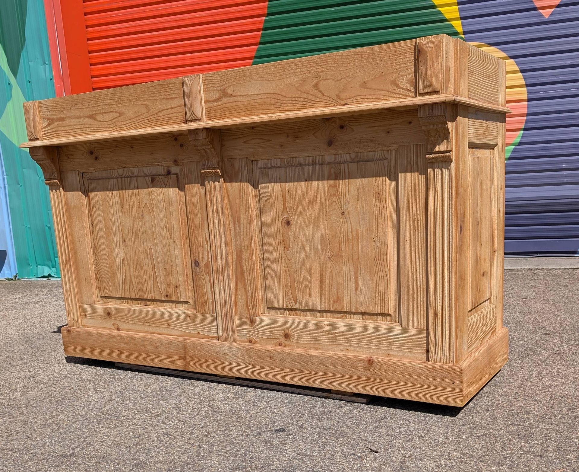 Bar en bois brun clair, installé à l'extérieur sur une surface gravillonnée, avec un mur peint de couleurs vives en arrière-plan