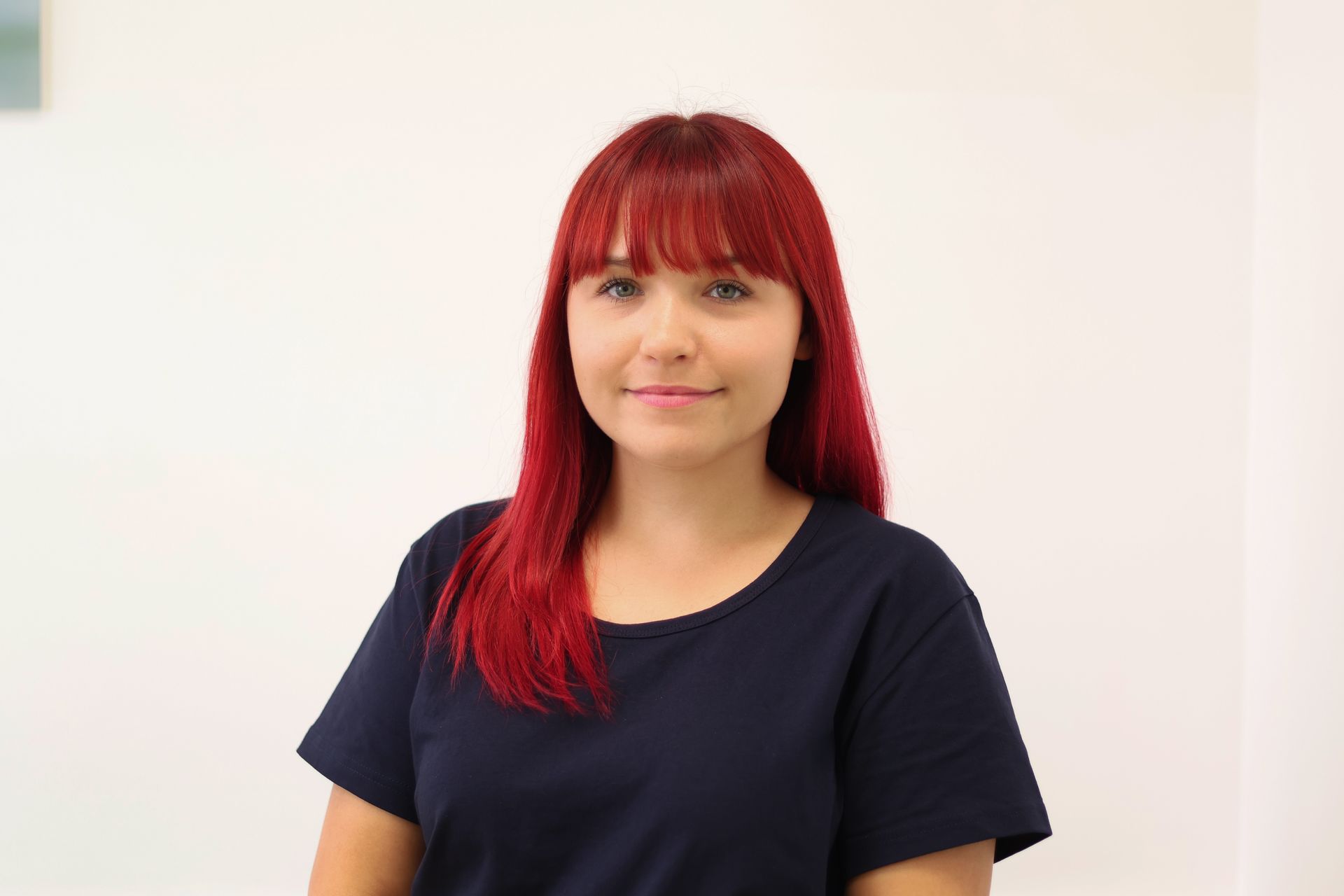 Frau mit roten Haaren und Pony in einem marineblauen Hemd, lächelt vor einem weißen Hintergrund in die Kamera.
