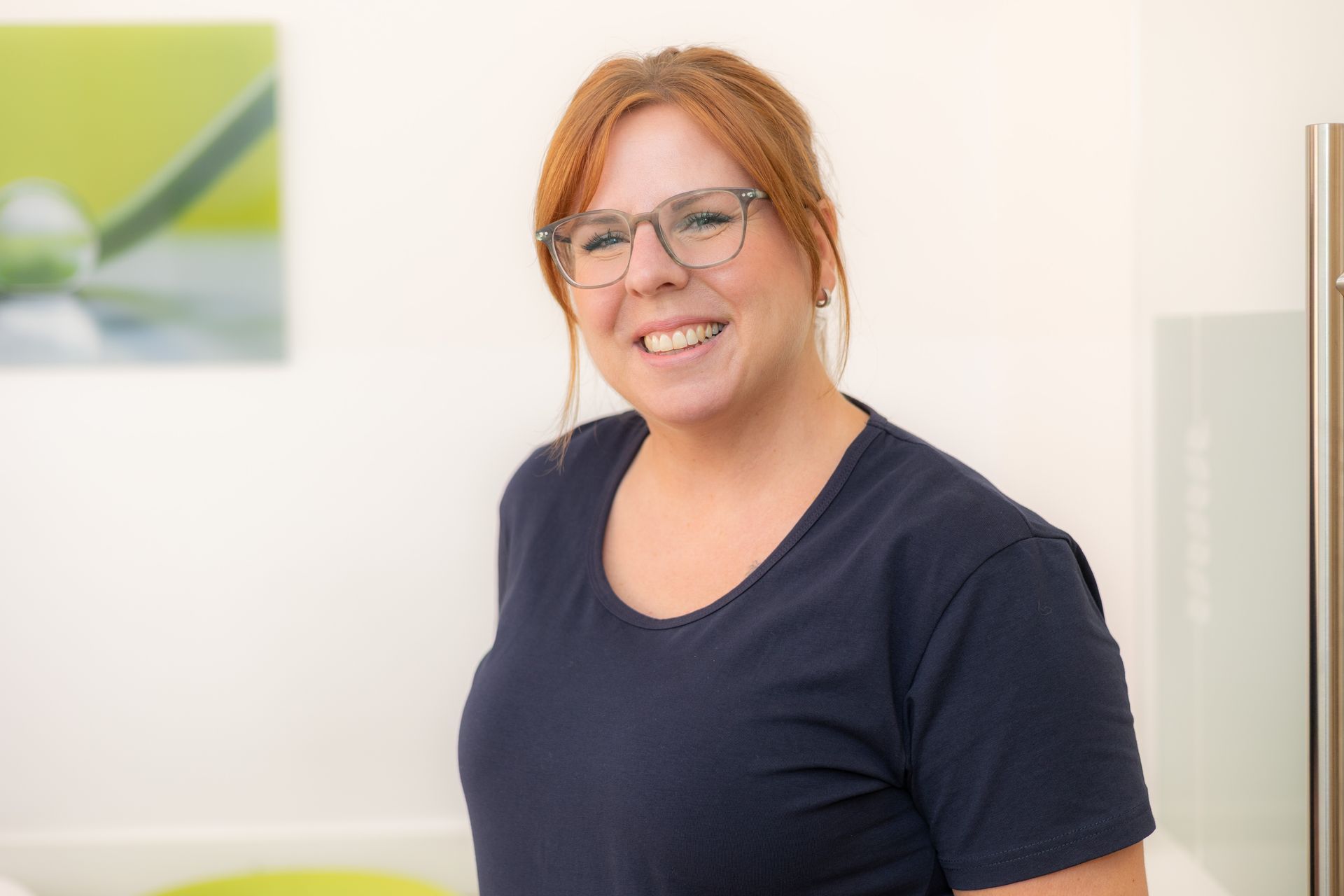 Frau mit roten Haaren und Brille lächelt, trägt ein blaues Hemd, in einem hellen Raum.