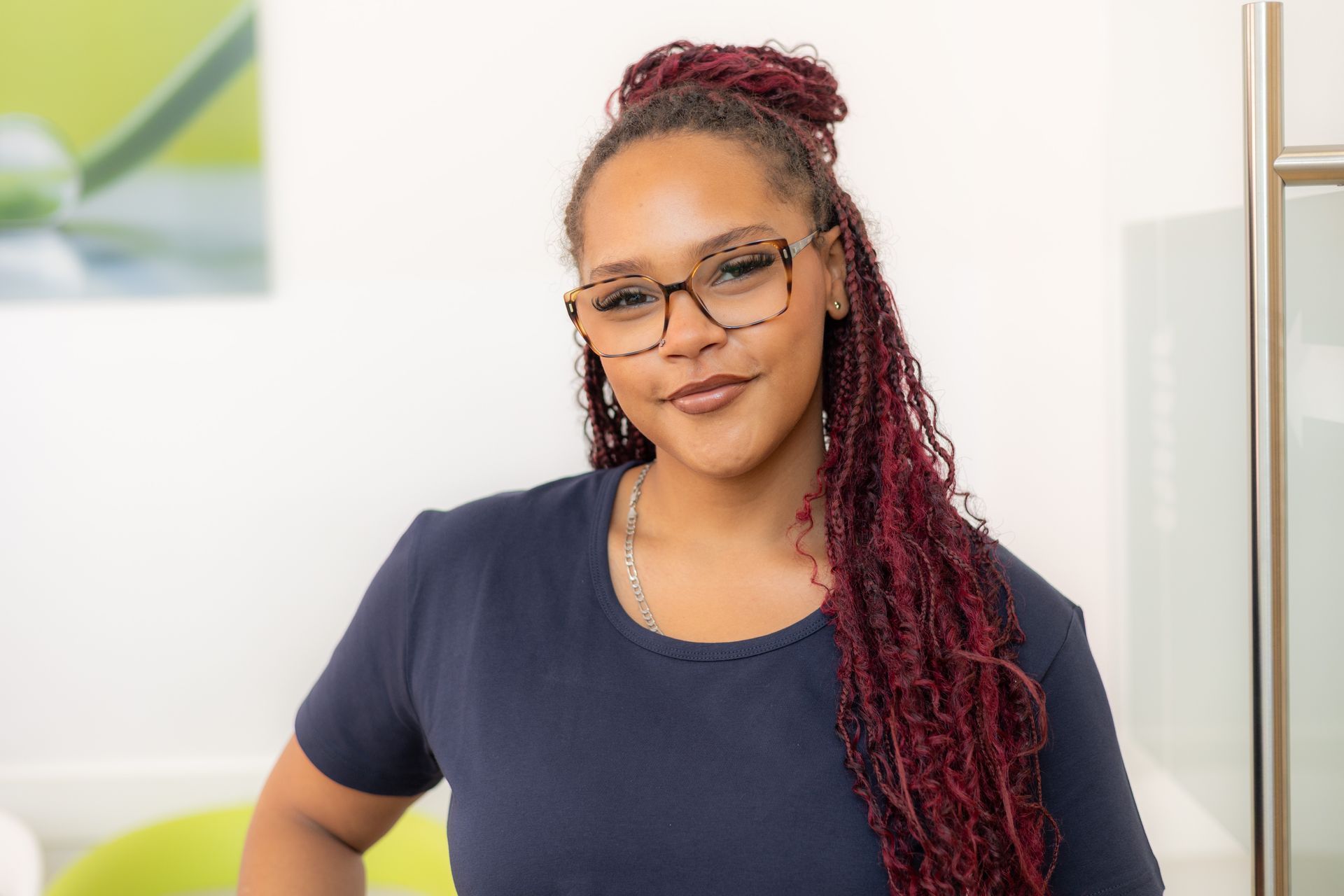 Frau mit roten Zöpfen, Brille und blauem Hemd lächelt. Drinnen, an weiße Wand gelehnt.