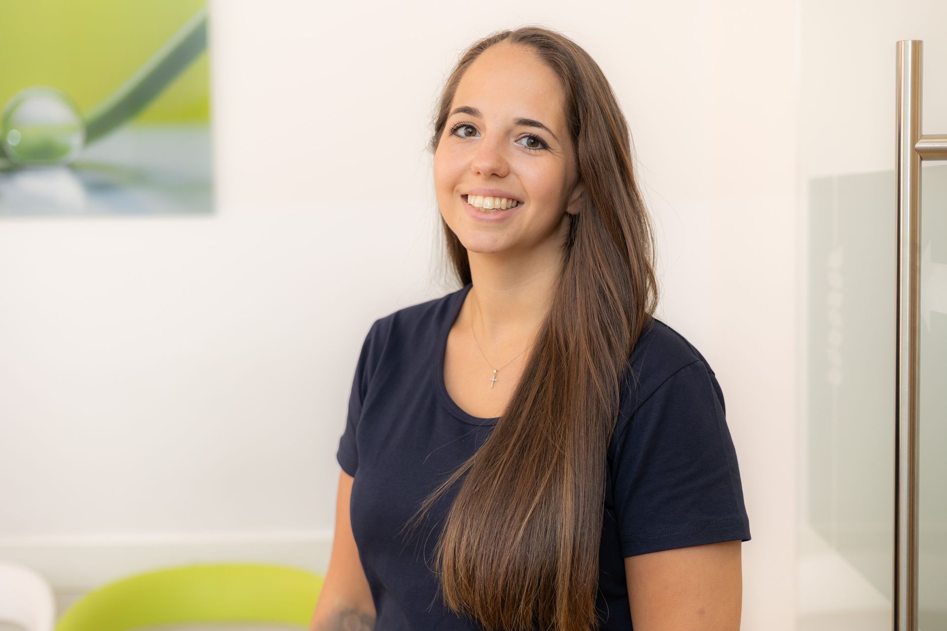 Frau mit langen braunen Haaren lächelt, trägt marineblaues Hemd, drinnen in der Nähe eines Kunstwerks.