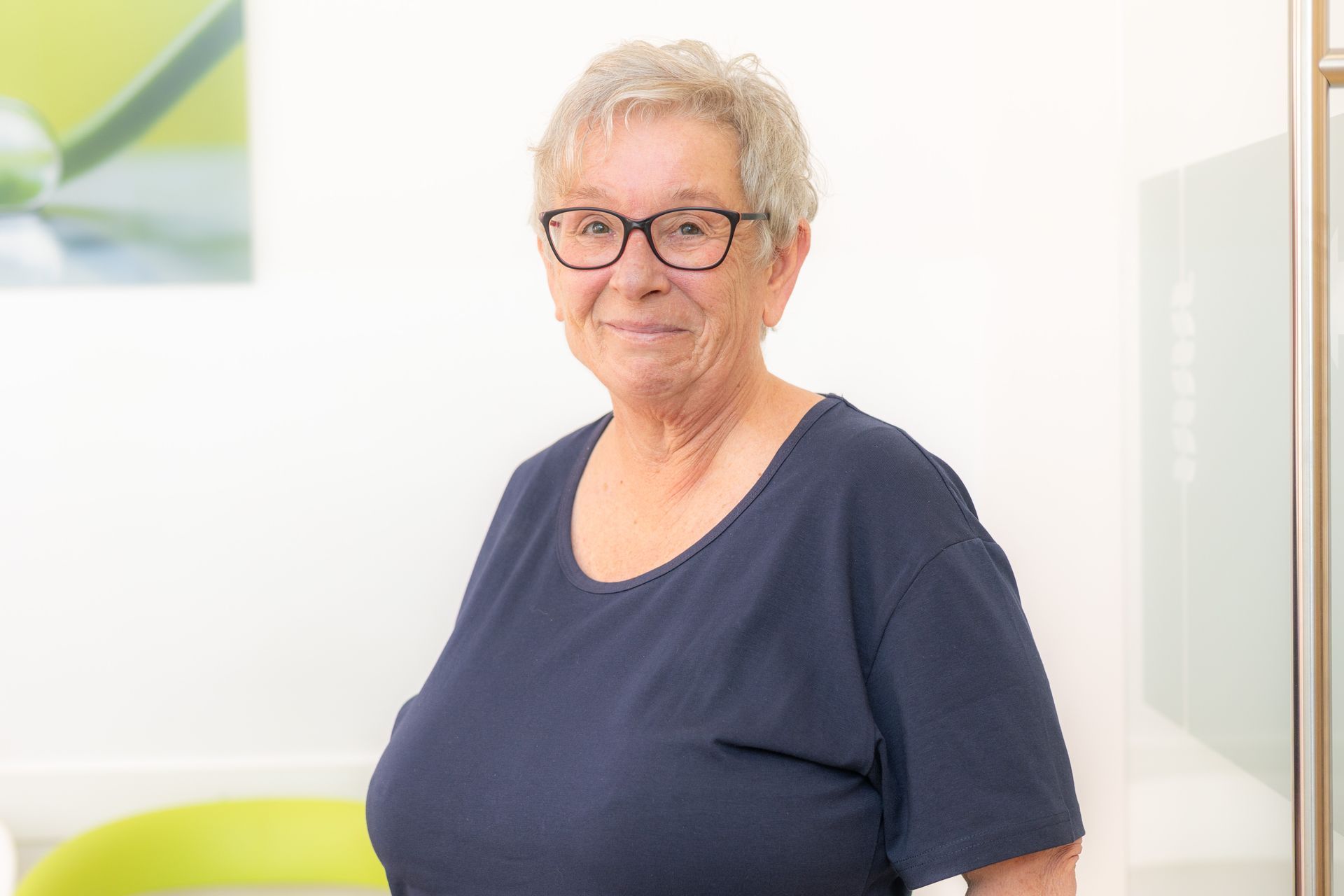 Eine ältere Frau mit Brille und blauem Hemd lächelt und steht im Haus.