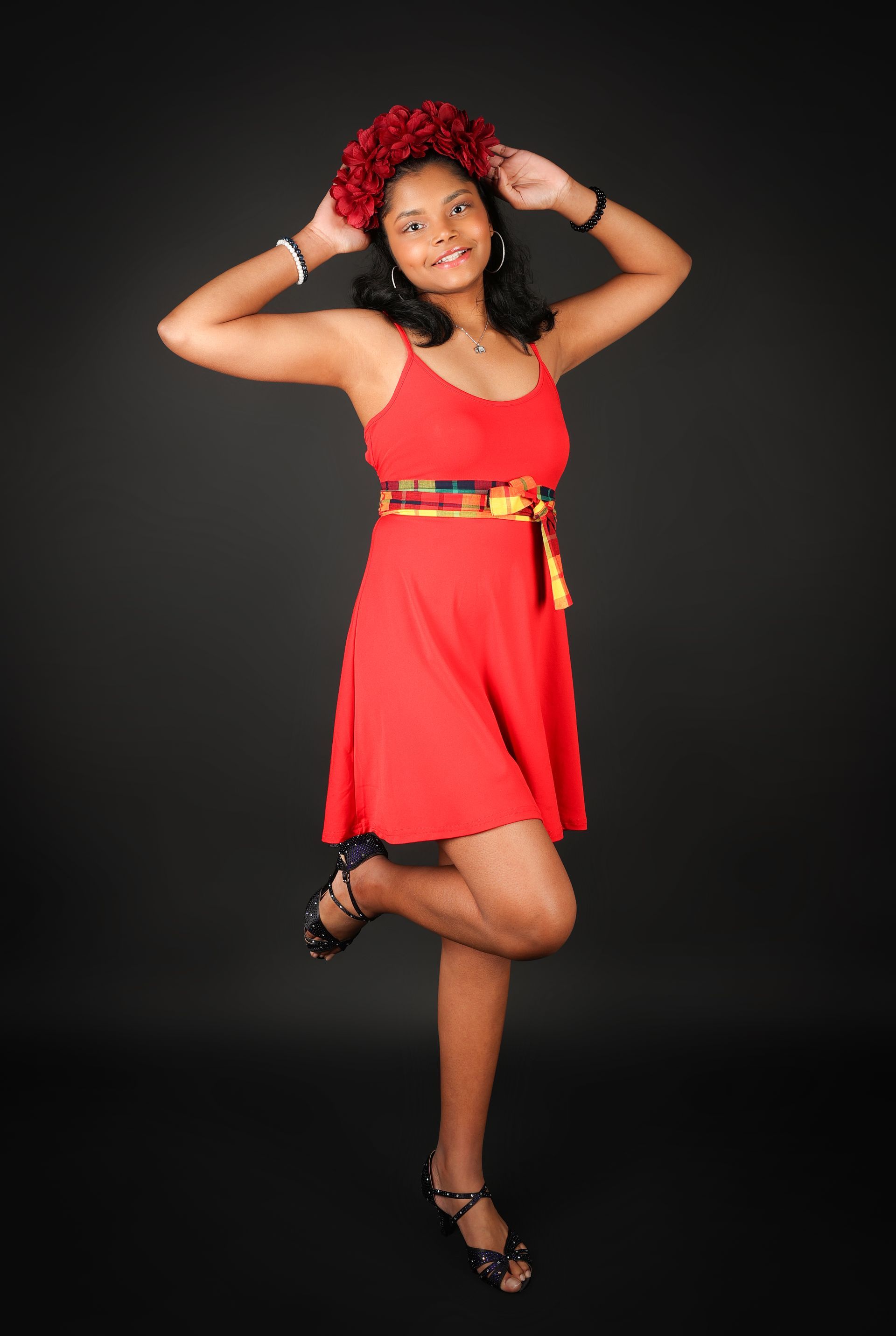 Femme en robe rouge posant sur fond noir, les mains sur la tête et un pied levé.