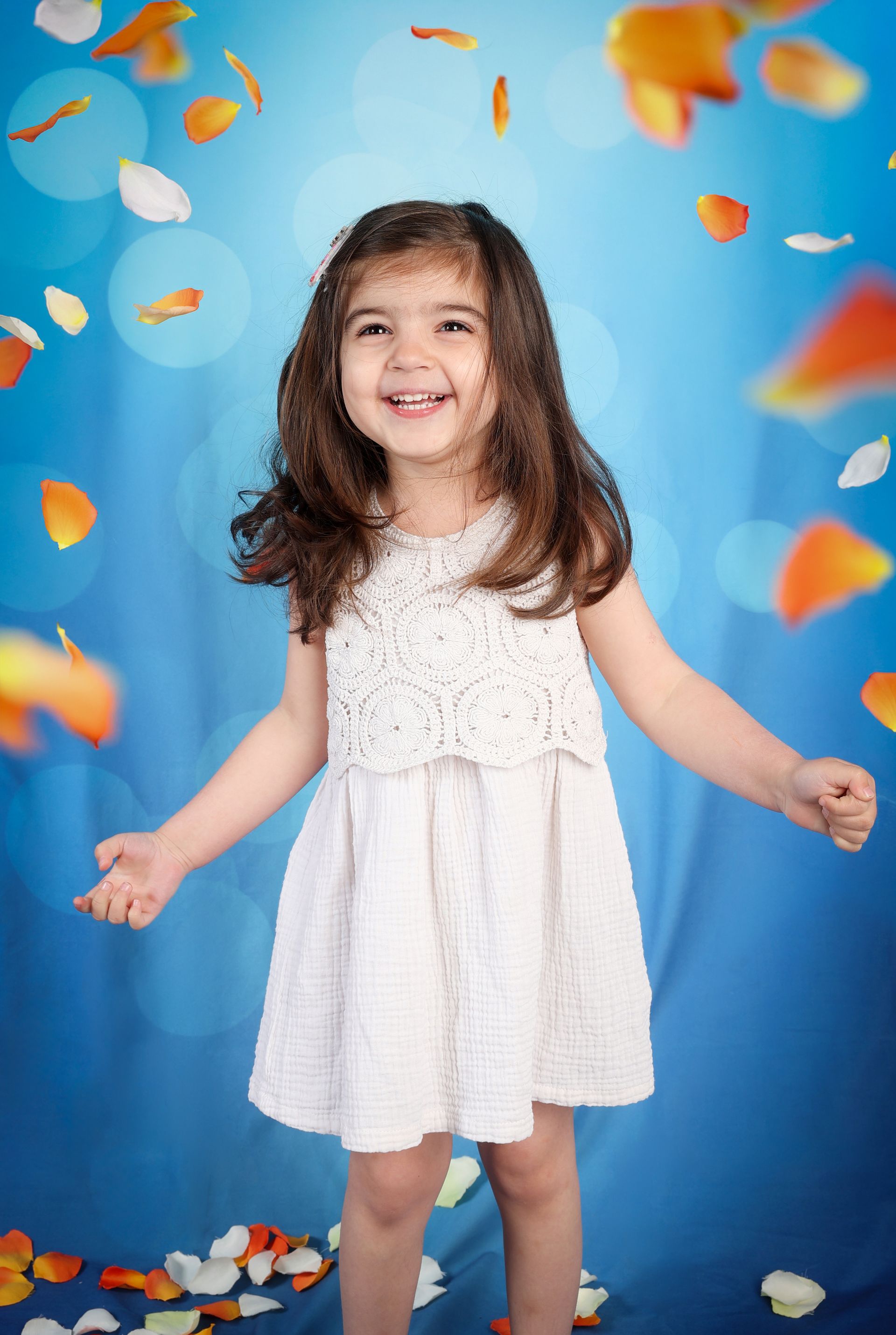 Enfant souriant en robe blanche sur fond bleu, avec des confettis colorés et des feuilles d’automne.