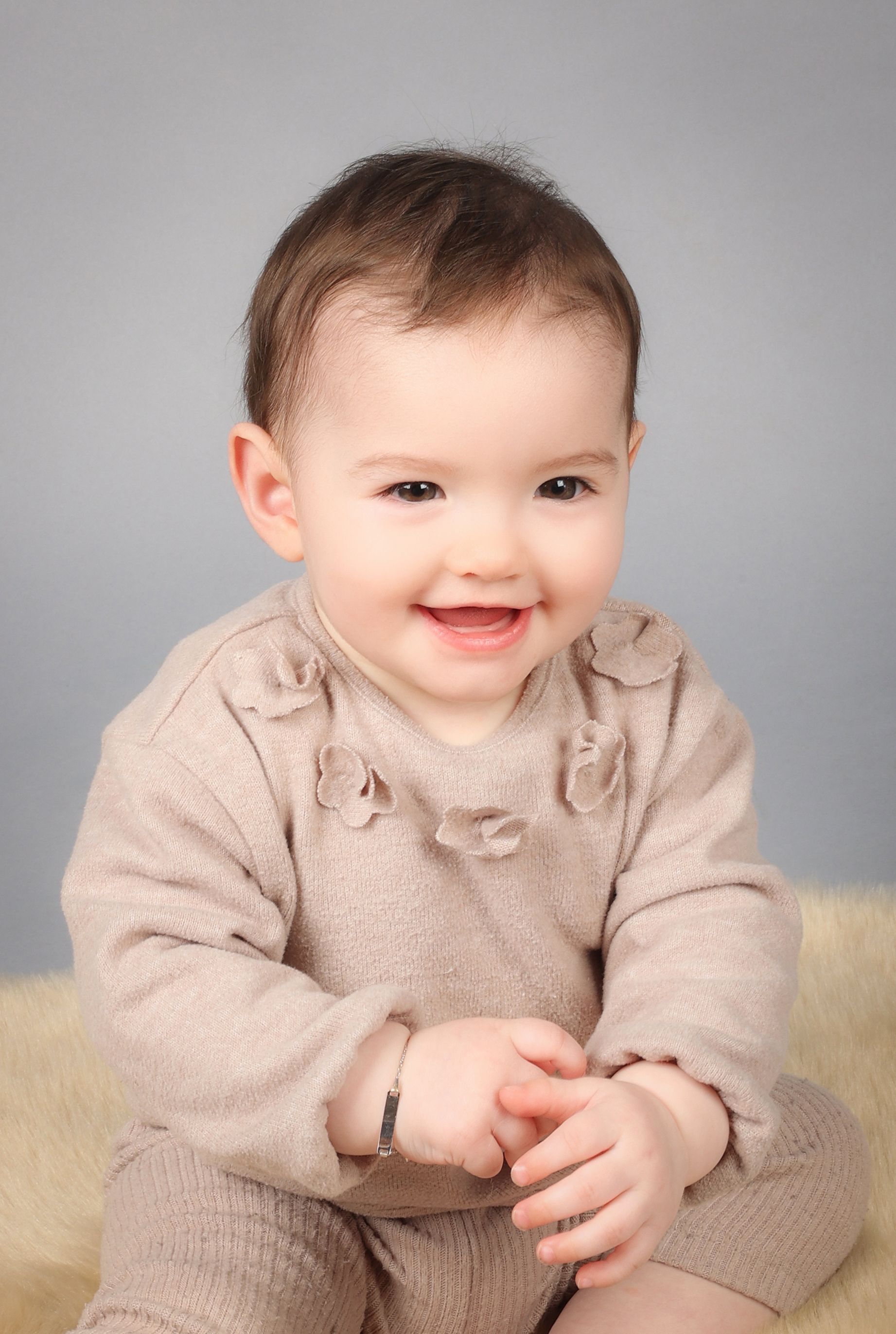 Bébé souriant assis sur un fond gris doux, vêtu d’une tenue beige et d’un bracelet en argent