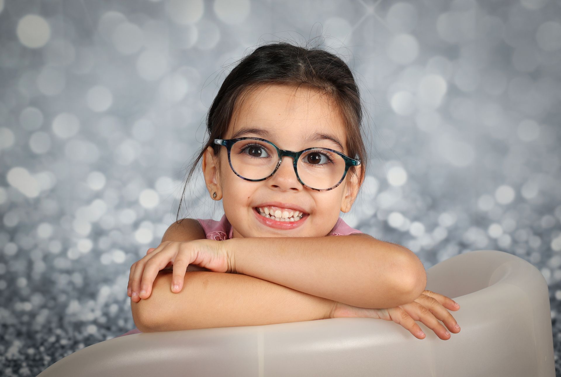 Enfant souriant avec des lunettes, appuyé sur ses bras croisés devant un fond gris flou à effet bokeh
