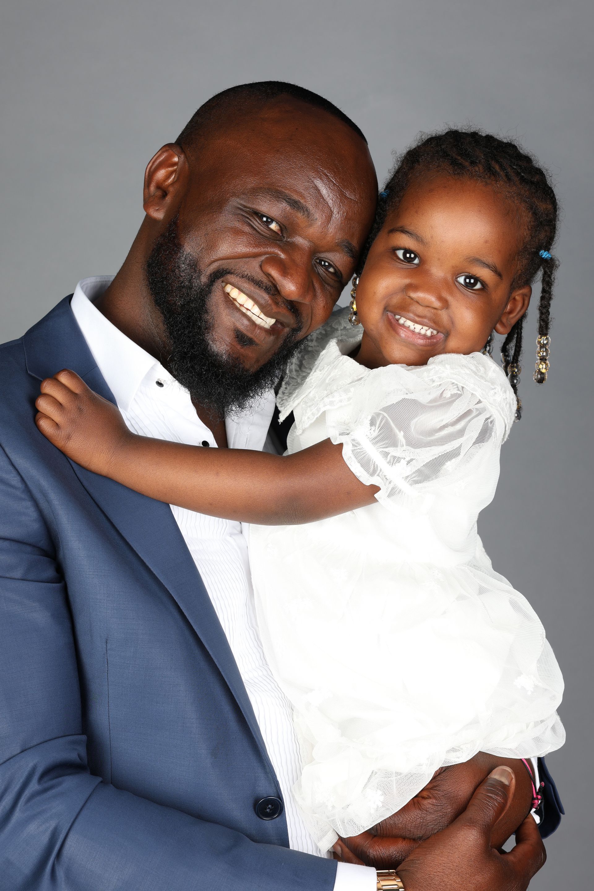 Homme souriant en costume bleu tenant une enfant en robe blanche sur fond gris