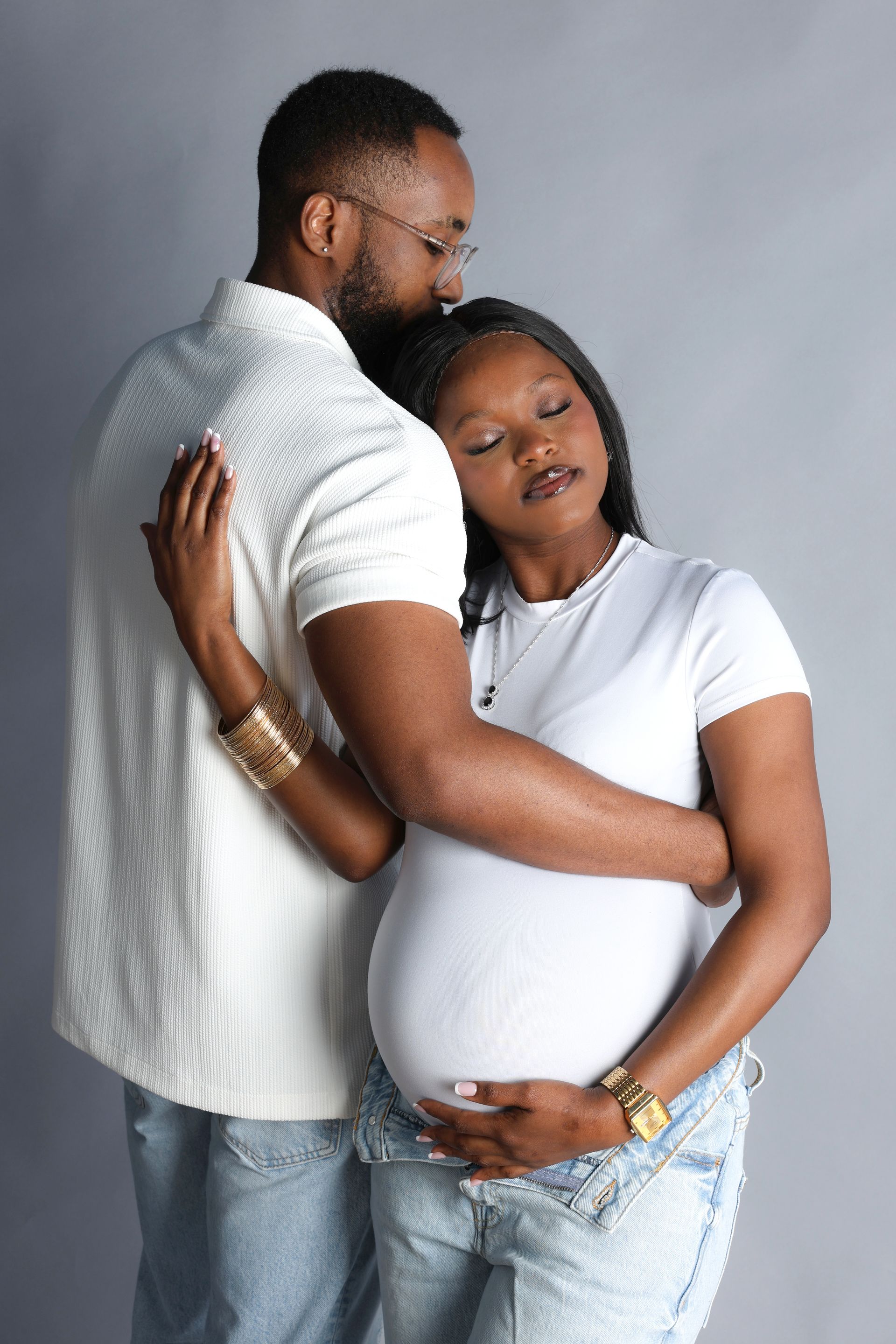 Un couple en chemises blanches s’enlace sur un fond de studio gris, l’un posant tendrement la main sur le ventre arrondi de l’autre