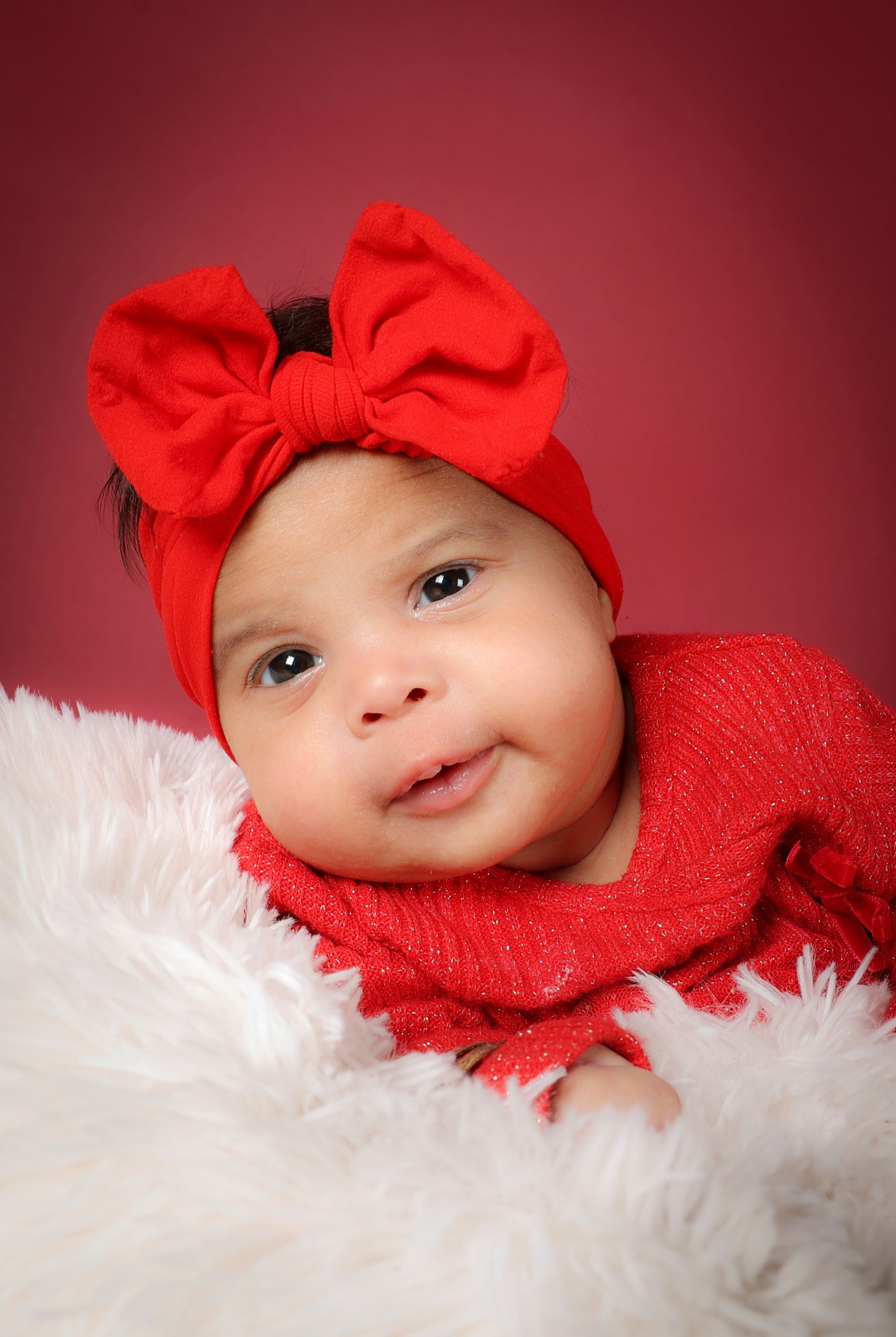 Bébé portant un bandeau rouge à nœud et une tenue rouge scintillante, posé devant un fond rose avec de la fourrure blanche.