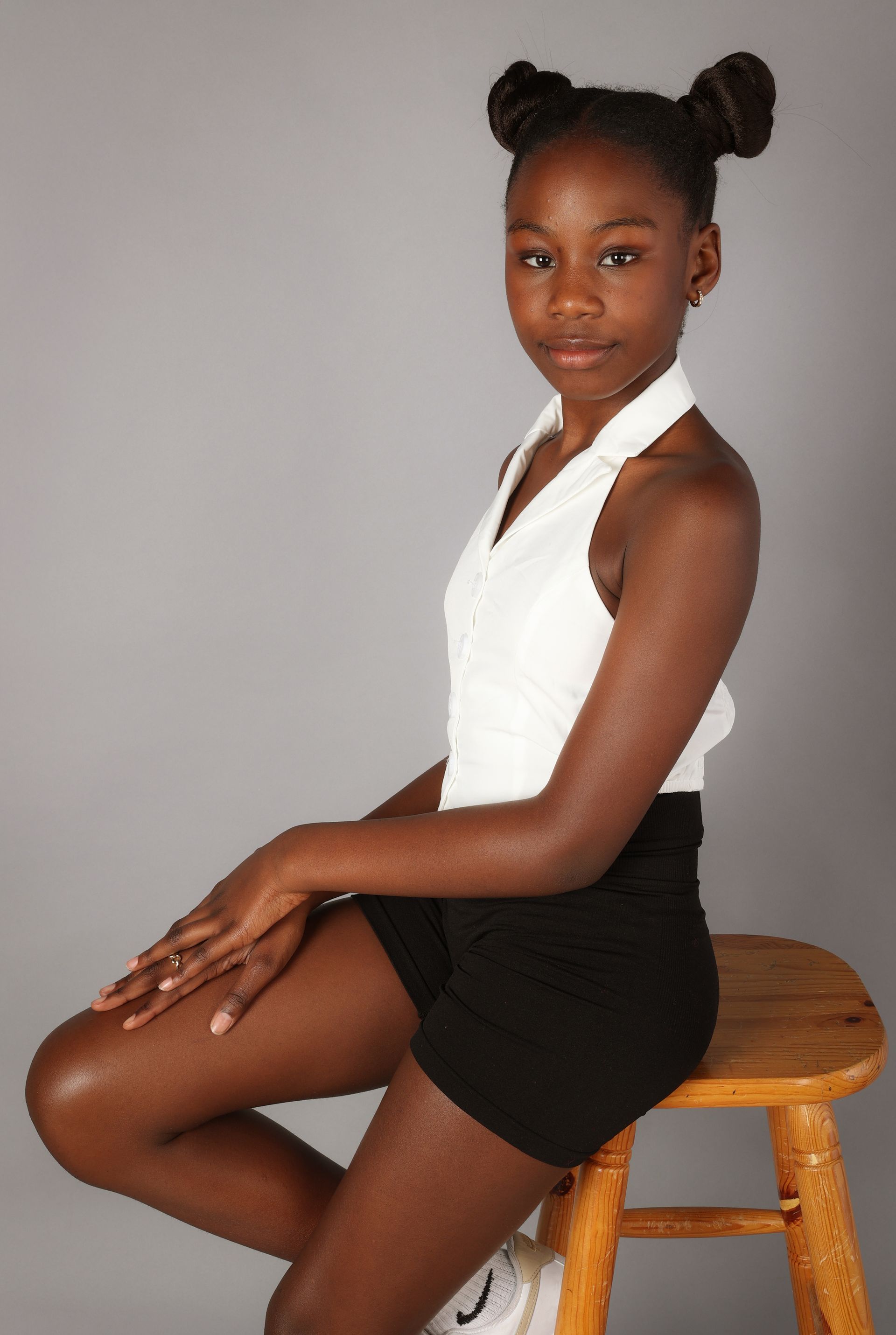 Personne assise sur un tabouret en bois, portant un haut blanc à dos nu et un short noir, devant un fond gris.