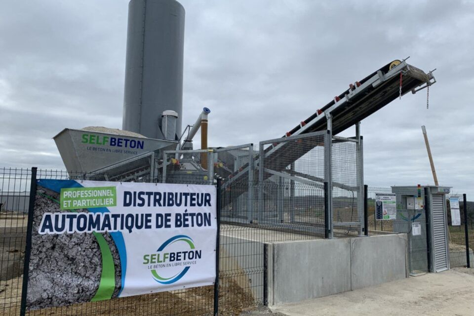 Une machine automatique de distribution de béton avec un grand silo et un tapis roulant derrière un panneau indiquant Selfbeton.