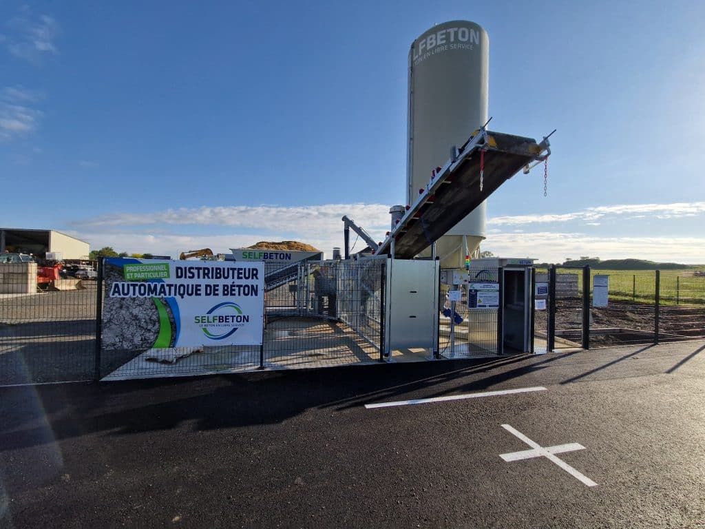 Une station de distribution de béton automatisée avec un haut silo et un tapis roulant, installée sur un terrain extérieur.