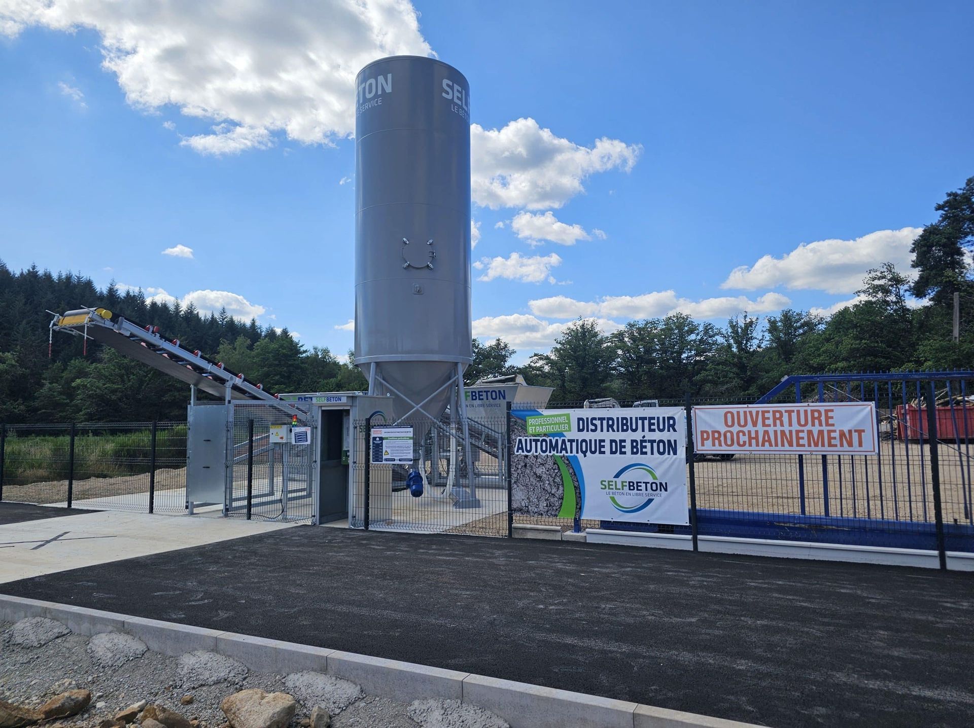 Un grand silo gris se dresse sur une esplanade extérieure pavée, avec des équipements industriels et des banderoles annonçant une ouverture future.