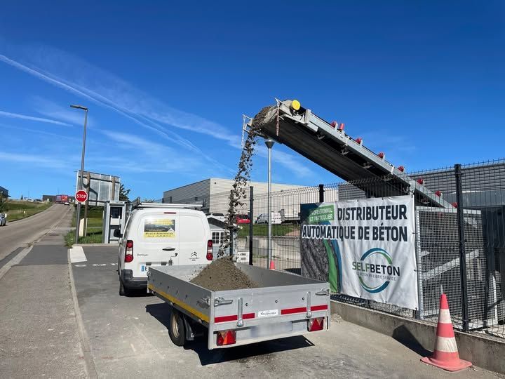 Une camionnette blanche avec une remorque se gare sous un convoyeur distribuant du béton dans une station de distribution automatisée de béton.