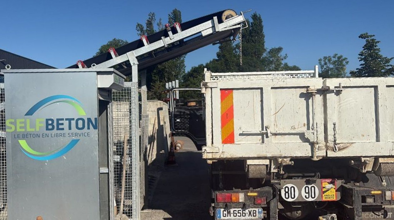 Une machine automatisée chargeant du béton à l'arrière d'un camion-benne sur un chantier extérieur.