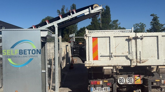 Une machine automatisée chargeant du béton à l'arrière d'un camion-benne sur un chantier extérieur.