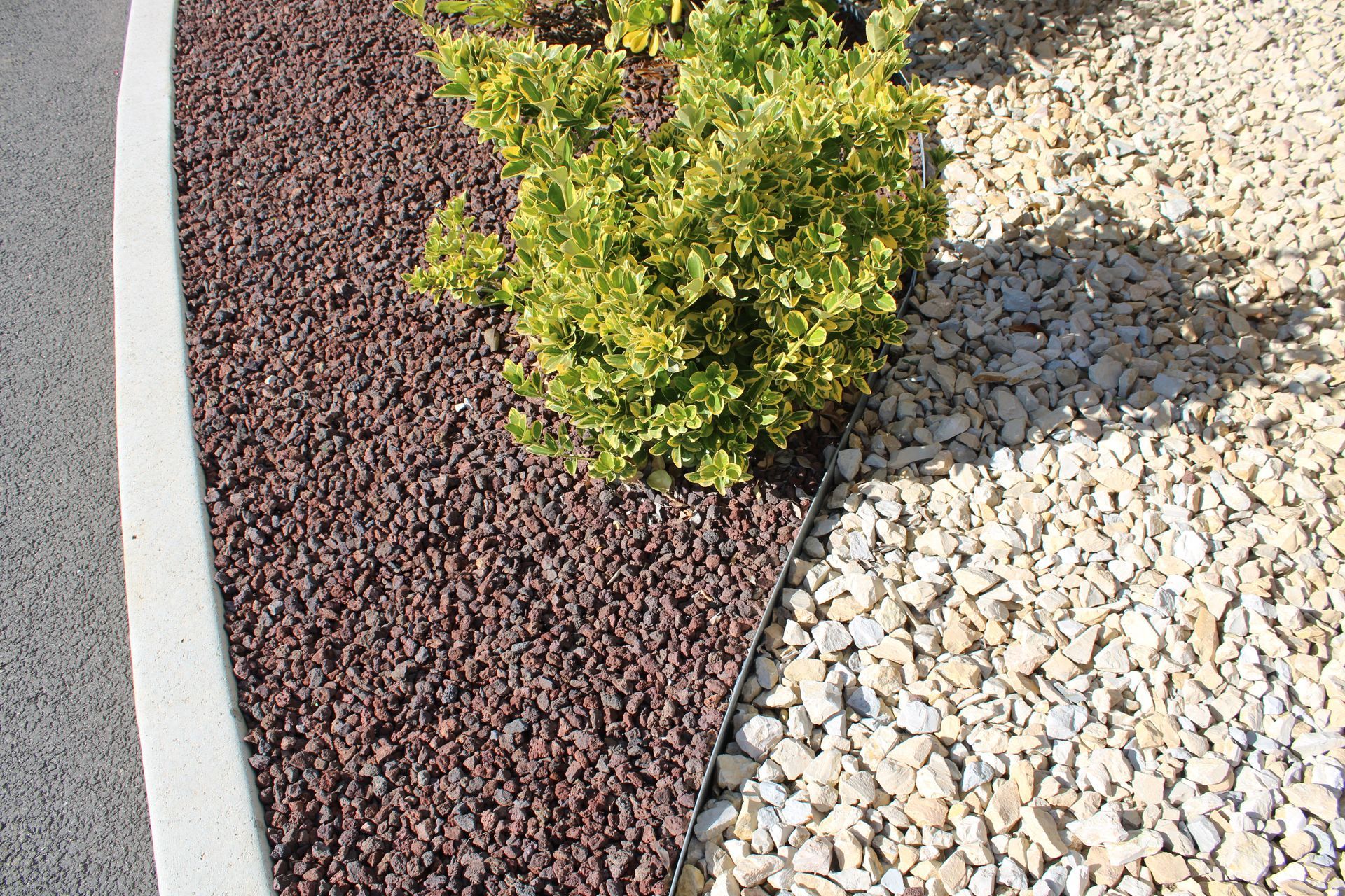 Un parterre de jardin incurvé est agrémenté de pierres concassées rouges, d'un petit buisson vert et d'une bordure de gravier beige clair près d'un trottoir.