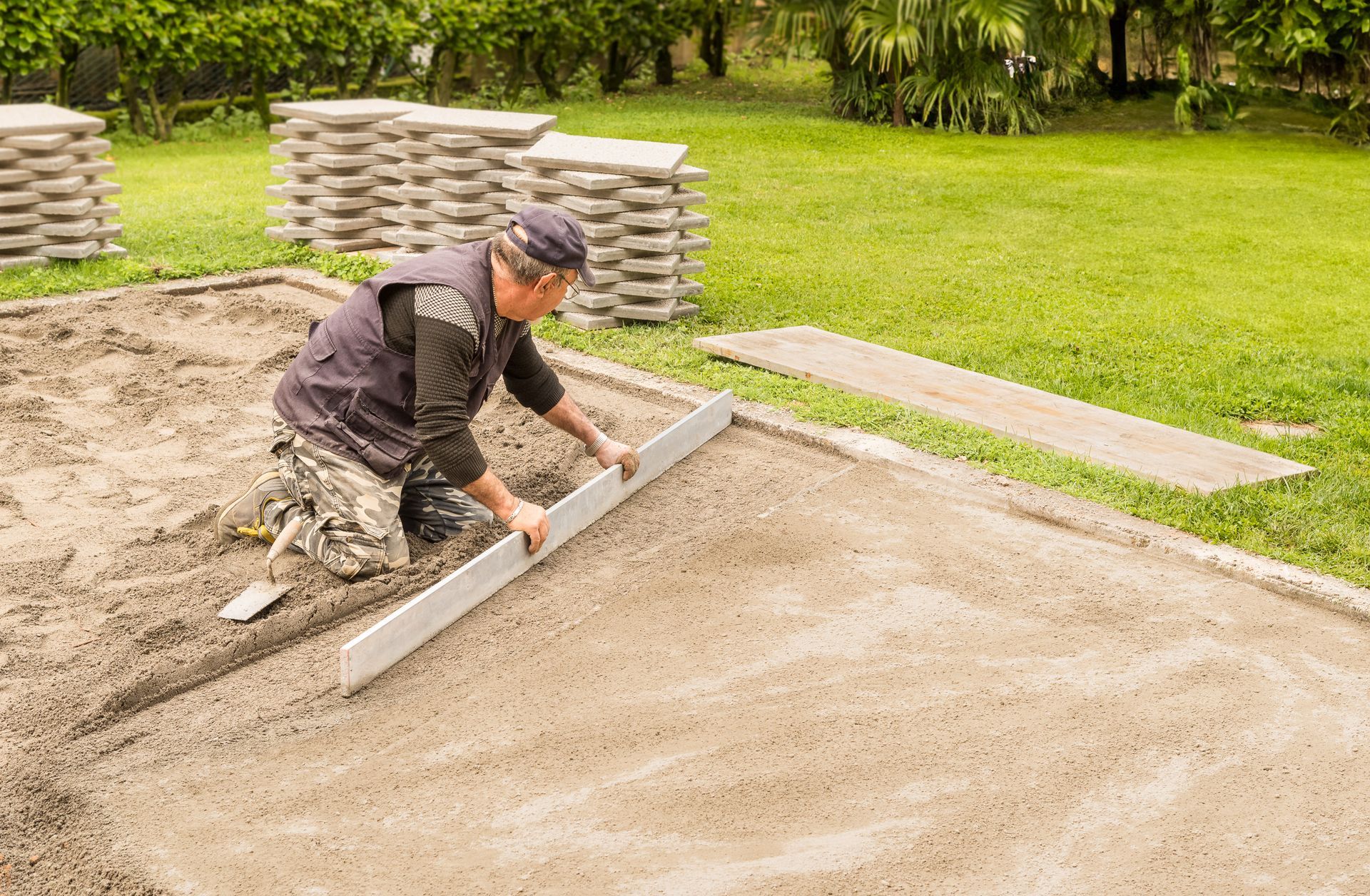 Une personne nivelle du sable avec une règle dans un jardin, préparant le terrain pour la pose de pavés empilés à l'arrière-plan.