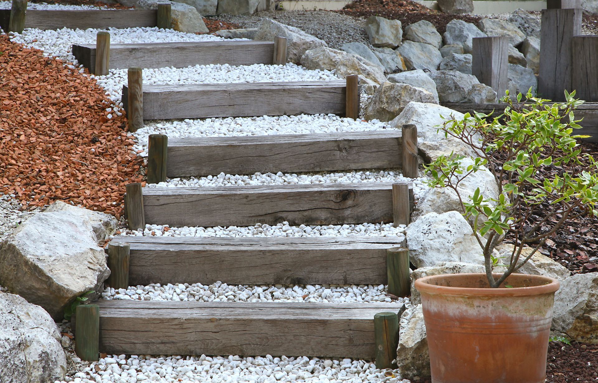 Un escalier de jardin en pierre et en bois recouvert de gravier blanc, flanqué de gros rochers, de copeaux de bois et d'une plante en pot.
