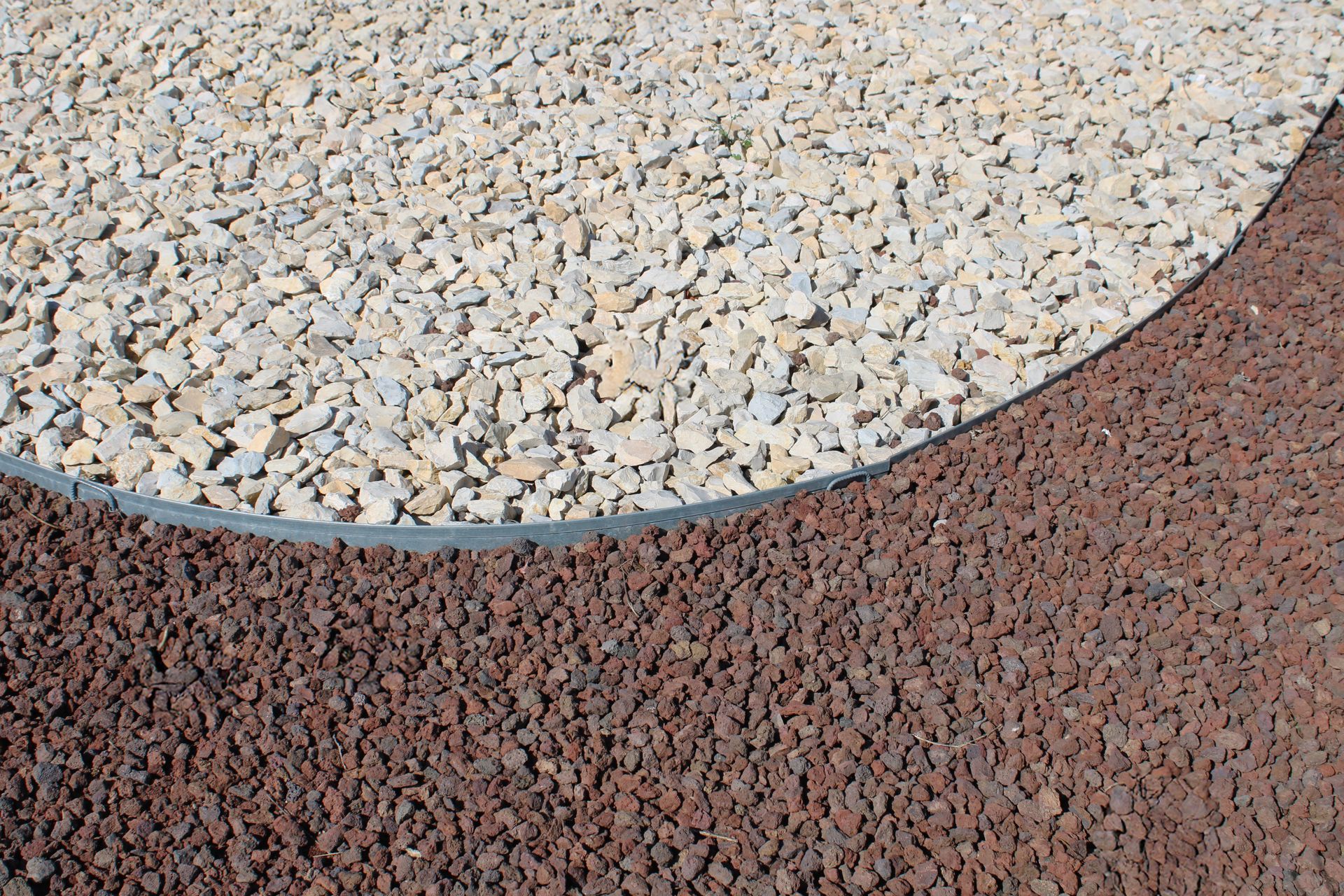 Vue de dessus montrant un rebord métallique incurvé séparant du gravier beige clair d'une zone de roche volcanique rouge foncé.